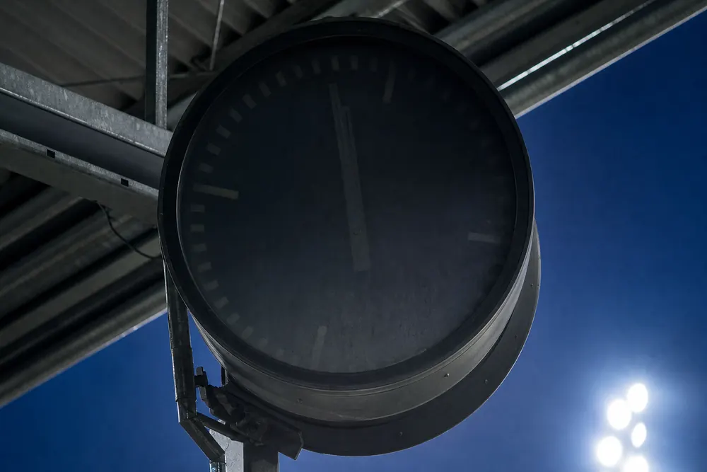 Große Stadionuhr unter dem Tribünendach, daneben ein Flutlichtmast im blauen Abendhimmel