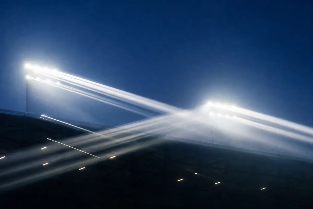 Zwei Flutlichtmasten leuchten im Nebel über ein nächtliches Stadion, die Lichtkegel kreuzen sich