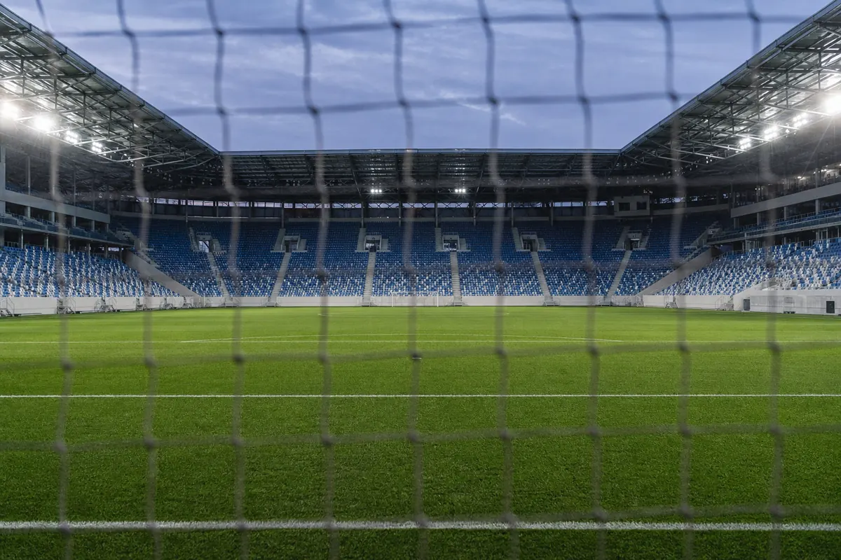 Blick durch das Tornetz auf ein leeres Stadion mit blauen Sitzschalen in der Dämmerung