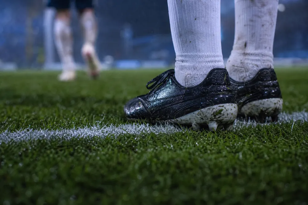 Nahaufnahme nasser Fußballschuhe an der Seitenlinie, im Hintergrund ein unscharfer Spieler