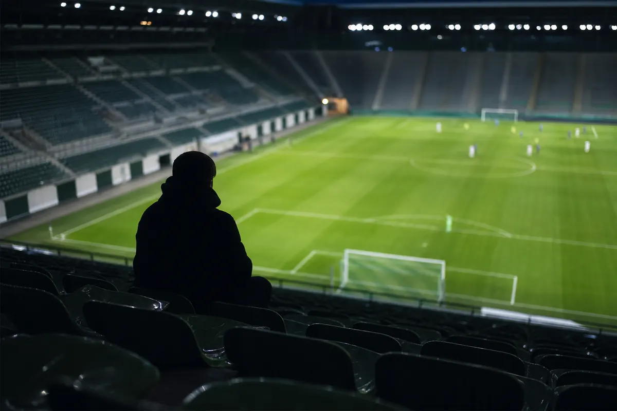 Ein einzelner Zuschauer sitzt auf der leeren Tribüne und blickt auf das beleuchtete Spielfeld